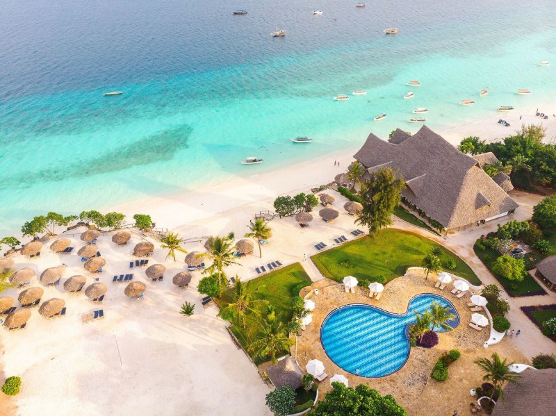Luftaufnahme eines luxuriösen Strandresorts mit herzförmigem Pool, strohbedeckten Sonnenschirmen, einem großen, mit Stroh gedeckten Gebäude und klarem, türkisfarbenem Meer mit Booten.