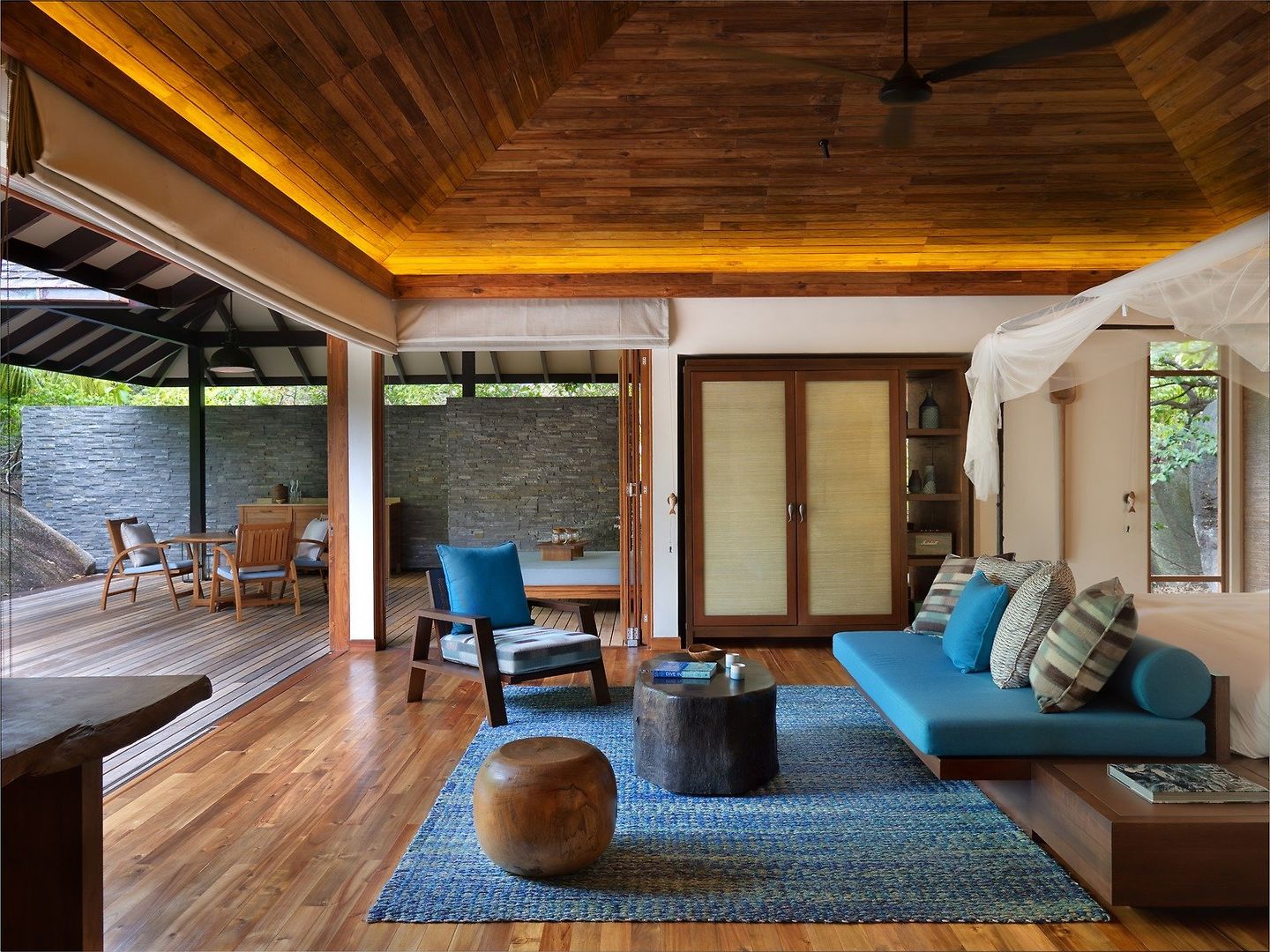 Wohnbereich einer Hideaway Pool Villa Ein geräumiges, modernes Wohnzimmer mit hohen Decken und Holzverkleidung. Eine Sitzgruppe mit türkisfarbenem Sofa und Sesseln steht auf einem bunten Teppich, daneben ein rustikaler Holztisch. Gegenüber dem Sitzbereich erkennt man eine Schlafnische mit einem großen Bett, umrahmt von einem Moskitonetz. Der Raum öffnet sich zu einer Außenterrasse mit Gartenmöbeln und Blick ins Grüne. Das Design verbindet moderne und natürliche Elemente zu einer eleganten und gemütlichen Atmosphäre.