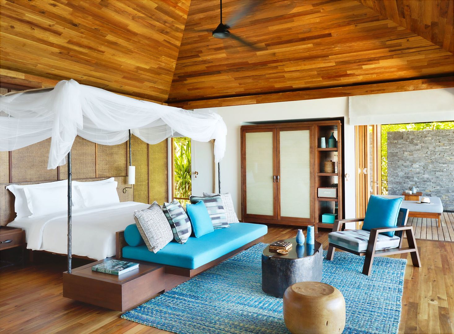 Hideaway Pool Villa Ein stilvoll eingerichtetes Schlafzimmer in einem Luxusresort mit einem großen Himmelbett, weißer Bettwäsche und einem Moskitonetz. Das Zimmer verfügt über holzgetäfelte Wände und eine Decke, eine blaue Sitzbank mit Kissen am Fußende des Bettes, einen Holzboden mit einem blauen Teppich und moderne Möbel. Ein großer Schrank, Sessel und Bücherregale ergänzen die Einrichtung. Große Fenster öffnen sich zu einem Außenbereich mit Naturblick.