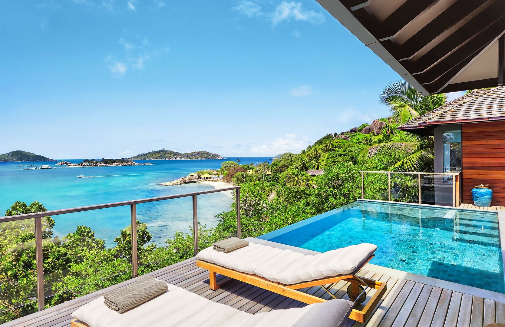 Ausblick von einer Oceanfront Pool Villa Blick von einer luxuriösen Terrasse mit einem Infinity-Pool über das türkisfarbene Meer und grüne Inseln. Zwei Sonnenliegen stehen auf dem Holzdeck, das von tropischer Vegetation umgeben ist, neben einem Teil eines Holzgebäudes mit Schiebetür. Ein klarer blauer Himmel überspannt die Szene.