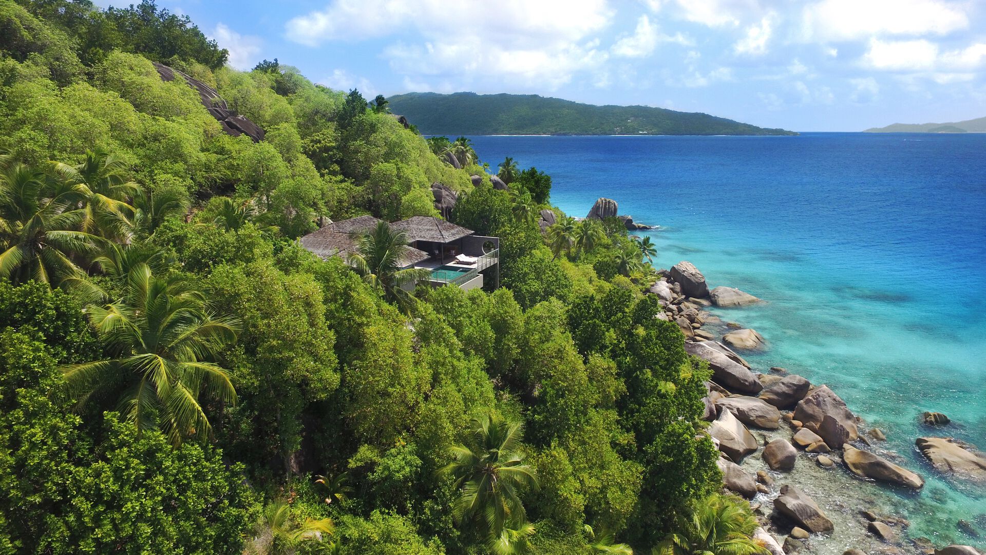 Oceanfront Pool Villa Ein luxuriöses Haus versteckt im dichten Grün eines tropischen Waldes an einem Hügel, mit Blick auf ein ruhiges türkisfarbenes Meer. Zahlreiche Palmen und andere Lush Vegetation umgeben das Anwesen, welches sich nahe der Küstenlinie mit großen Felsen am Ufer befindet.