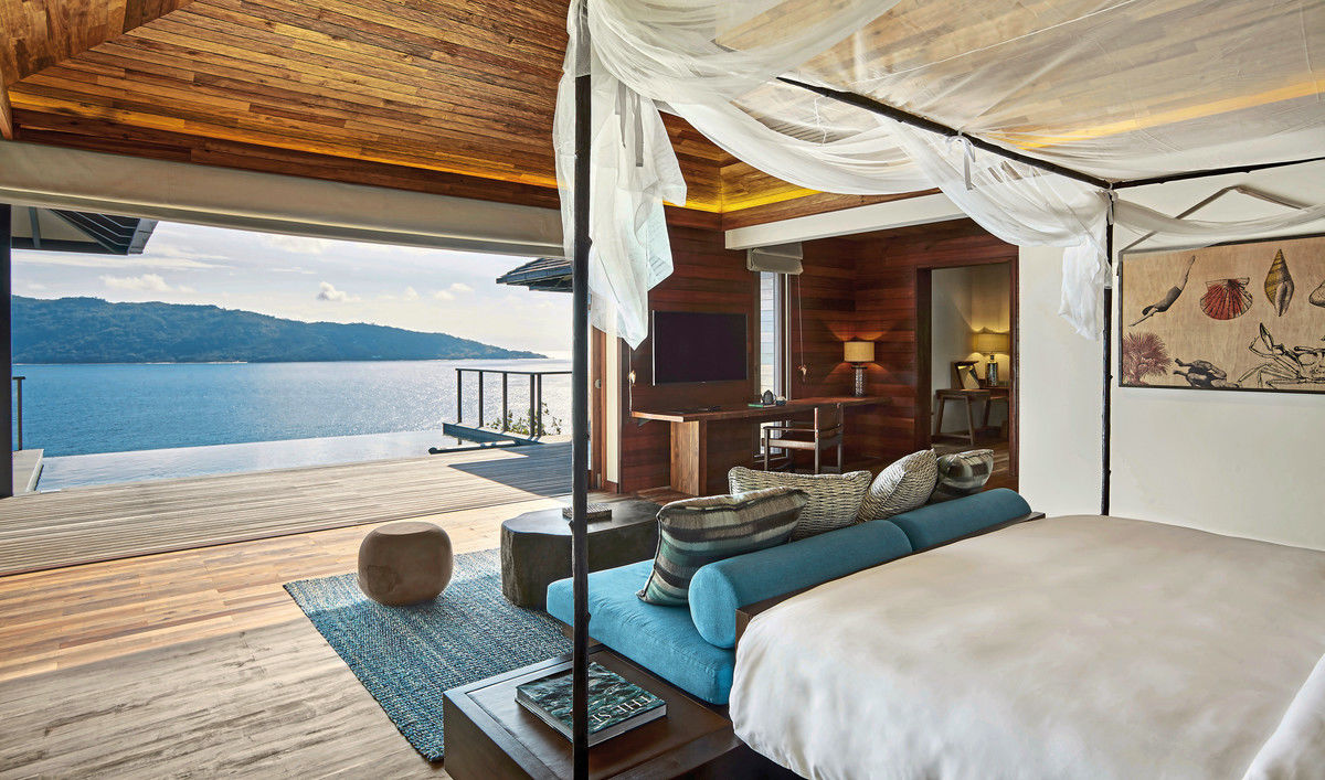 Ausblick von einer Panorama Pool Villa Ein luxuriöses Schlafzimmer mit Blick auf das Meer, ausgestattet mit einem großen Bett mit weißer Bettwäsche und einem luftigen Moskitonetz. Holzpaneele schmücken die Wände und Decke, während im Hintergrund eine offene Terrasse mit atemberaubender Aussicht zu sehen ist. Dekorative Kissen und ein Wandteppich mit Meeresmotiv verleihen dem Raum eine gemütliche Atmosphäre.