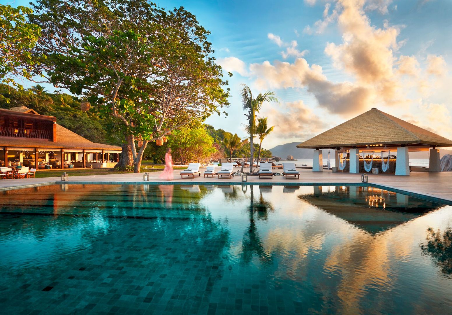Pool Ein luxuriöser Infinity-Pool mit klarem blauen Wasser vor einem exotischen Resort. Im Hintergrund befindet sich ein traditionelles Gebäude mit Strohdach und eine Reihe von Sonnenliegen unter Palmen. Der Himmel zeigt leichte Wolken und leuchtet in warmen Tönen der untergehenden Sonne. Ein friedvolles Paradies, das zum Entspannen einlädt.
