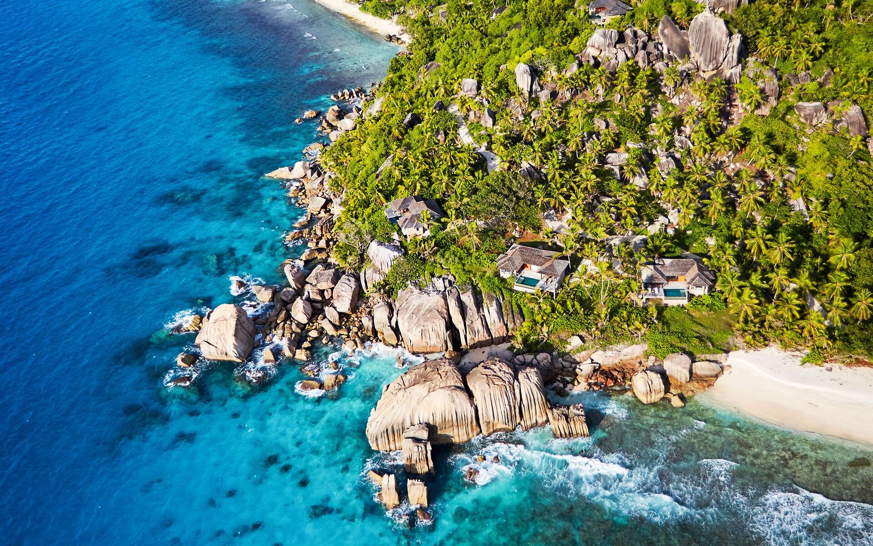Blick auf das Six Senses Zil Pasyon Luftaufnahme einer tropischen Inselküste mit üppiger Vegetation, Felsformationen und einigen verstreuten Bungalows am Meer, umgeben von klarem, blauem Wasser.