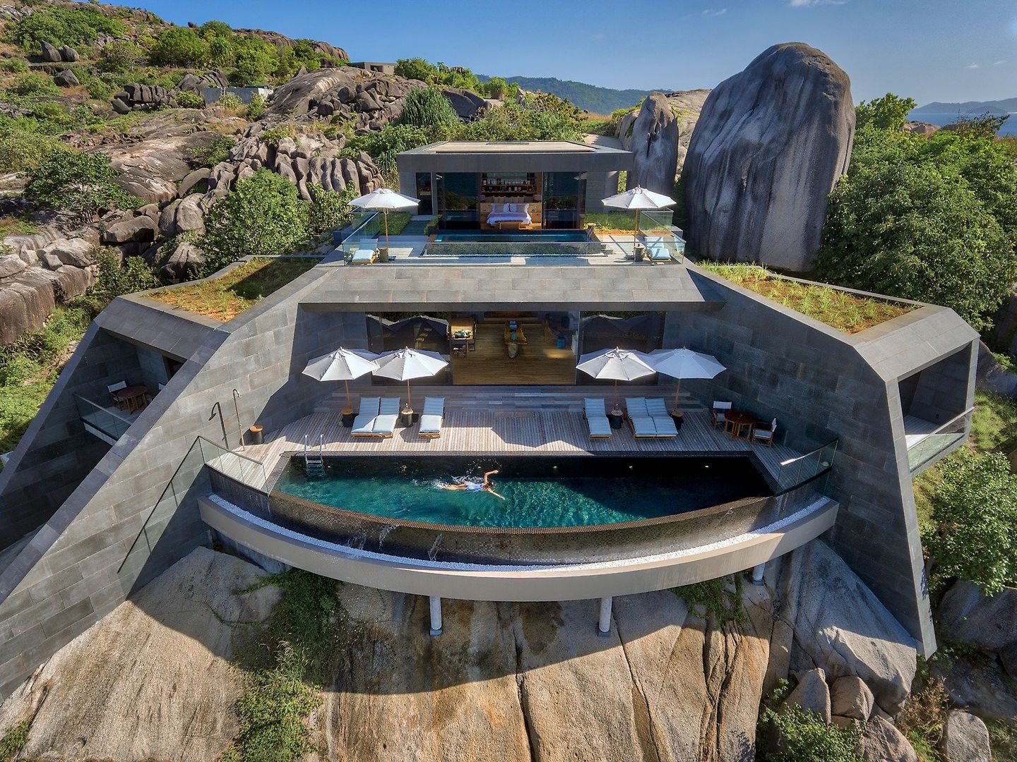 Seascape Three Bedroom Residence Eine moderne Villa, eingebettet in eine felsige Landschaft mit üppiger Vegetation. Die Architektur zeigt klare Linien und offene Wohnbereiche, die sich auf mehrere Ebenen erstrecken, mit einem zentral platzierten Infinity-Pool. Sonnenliegen und schattenspendende Schirme befinden sich auf einer Terrasse neben dem Pool. Eine Person schwimmt im Pool. Das Gebäude scheint harmonisch in die natürliche Umgebung integriert und bietet einen atemberaubenden Blick auf die umliegende Natur.