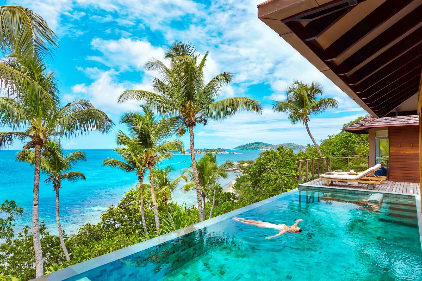 Sgnature Two Bedroom Pool Villa Ein Infinity-Pool mit einer Person, die darin schwimmt und Blick auf tropisches Meer mit Kokospalmen. Eine Sonnenliege befindet sich auf einer Holzterrasse neben einem modernen Gebäude.