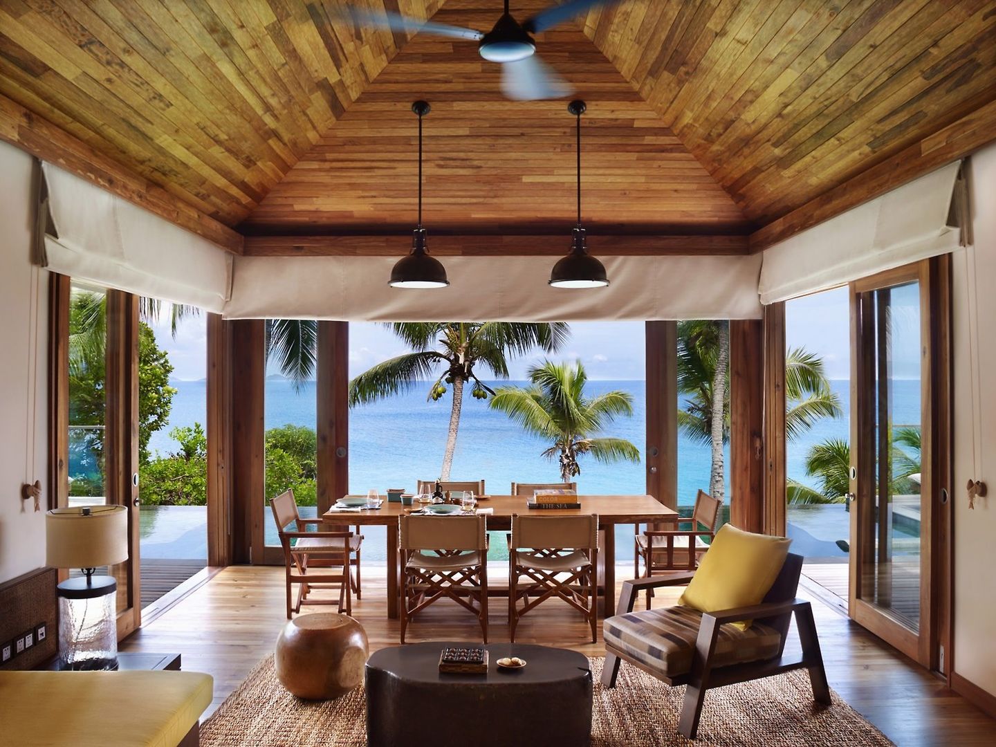 Wohnbereich in einer Signature Two Bedroom Pool Villa Innenaufnahme eines luxuriösen Strandhauses mit eleganten Holzmöbeln, einem Esszimmer, das sich zu einer Terrasse mit Meerblick öffnet, umgeben von tropischen Palmen.