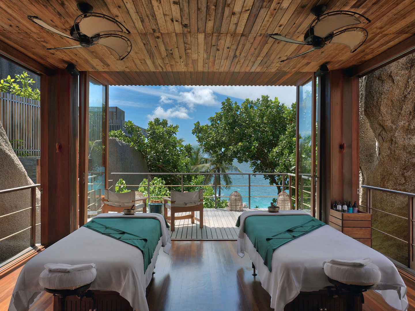 Spa Ein luxuriöses Spa-Zimmer mit Holzdecke und zwei Massagetischen, die zum geöffneten Balkon mit Blick auf das türkisfarbene Meer und tropische Vegetation hin ausgerichtet sind. Natürliches Licht durchflutet den Raum, verstärkt von den warmen Holztönen und der friedlichen, einladenden Atmosphäre.