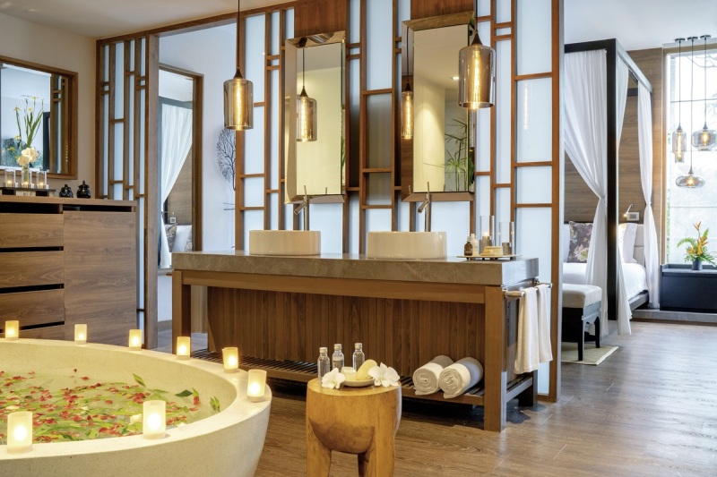 Badezimmerbeispiel Ein luxuriöses Badezimmer im Spa-Stil mit Doppelwaschbecken, großen Spiegeln, einer gefliesten Badewanne mit Blütenblättern, umgeben von Kerzen. Im Hintergrund ist der Eingang zu einer Entspannungslounge mit einem Massagebett zu erkennen. Die Einrichtung ist in warmen Brauntönen mit asiatisch inspirierten Designelementen gehalten.