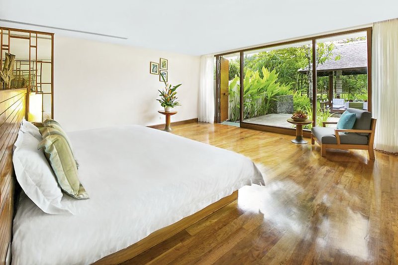 Garden Residence Ein geräumiges, modern eingerichtetes Schlafzimmer mit einem großen Bett mit weißer Bettwäsche, Holzfußboden und großen Fenstern, die den Blick auf einen üppigen Garten freigeben. Neben dem Bett befindet sich ein kleiner Sessel und ein Tisch mit dekorativen Elementen.