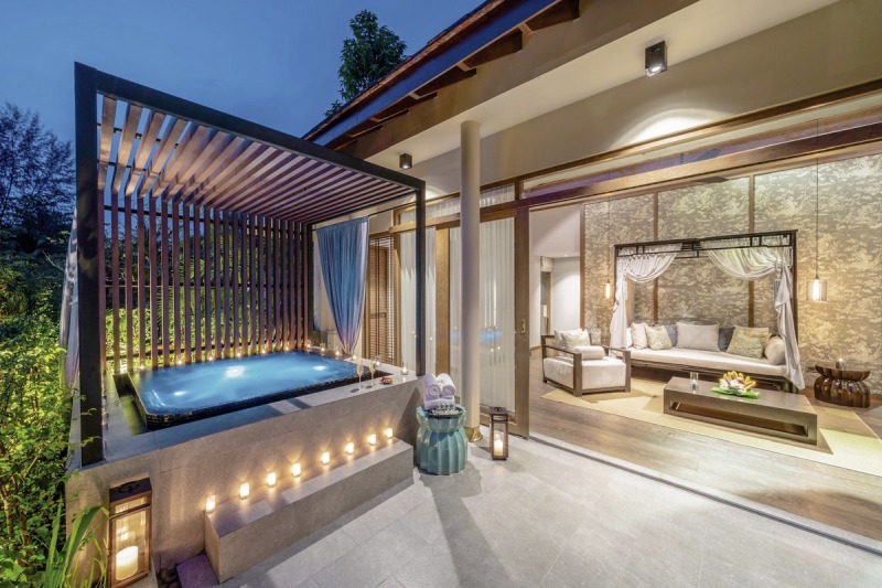 Jacuzzi Pool Suite Luxuriöse Außenterrasse bei Dämmerung mit Whirlpool, integriert in ein modernes Design mit hölzernem Sichtschutz und Beleuchtung. Im Hintergrund ist ein elegantes Wohnzimmer mit offenen Glasschiebetüren und stilvoller Inneneinrichtung zu sehen.