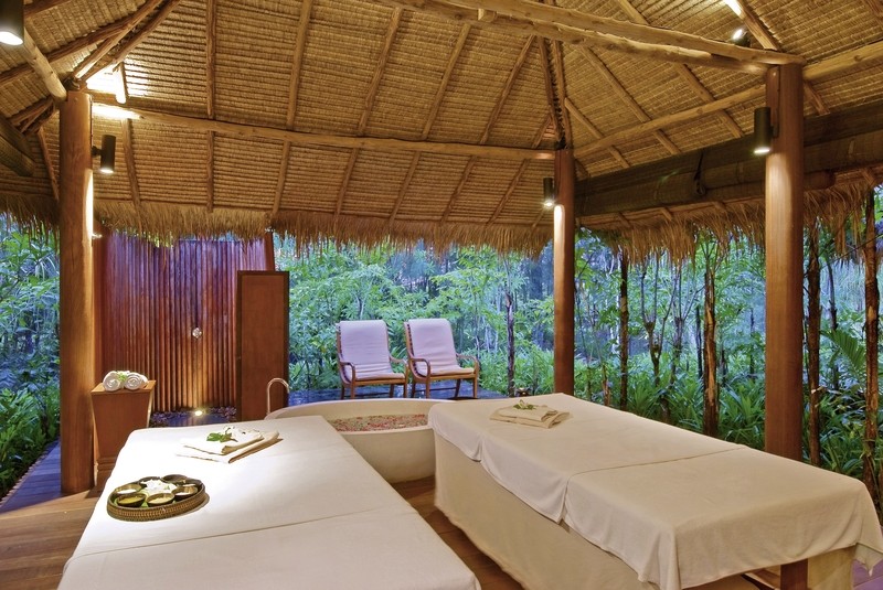 Massagepavillon Ein ruhiger Wellnessbereich im Freien mit zwei Massageliegen, die mit weißen Tüchern bezogen sind. Im Hintergrund befinden sich zwei gepolsterte Liegestühle auf einer Terrasse, die einen entspannten Blick auf einen üppigen, dicht bewachsenen Dschungel bieten. Die natürliche Atmosphäre wird durch ein traditionelles Strohdach und rustikale Holzelemente unterstrichen.