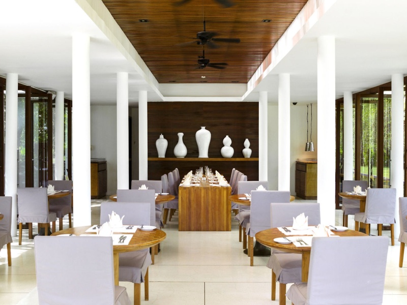 Restaurant Eine elegante, geräumige Esshalle mit weißen Säulen, Holzdecke und großen Vasen auf einem Sideboard im Hintergrund. Die Tische sind sorgfältig mit weißen Tischtüchern, Geschirr und gefalteten Servietten gedeckt. Große Fenster bieten einen Blick auf das umgebende Grün.
