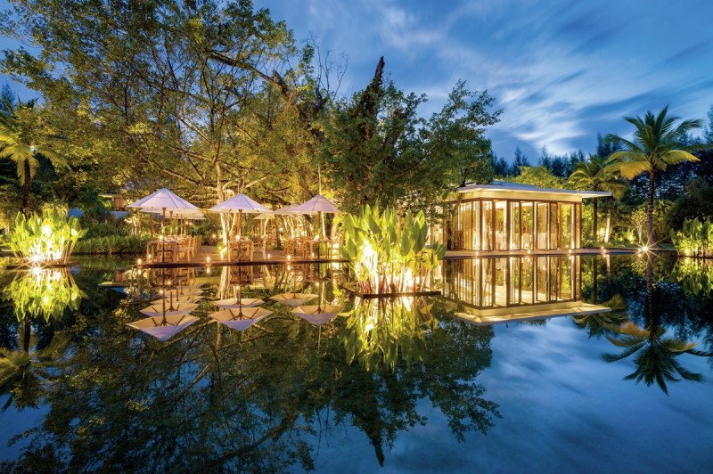 Restaurant Ficus Ein elegantes Außenrestaurant in der Abenddämmerung mit beleuchteten Tischen unter weißen Sonnenschirmen an einem ruhigen Teich. Im Hintergrund ein modern gestaltetes gläsernes Pavillonhaus, umgeben von tropischer Vegetation und einer Spiegelung der Szene auf der Wasseroberfläche.