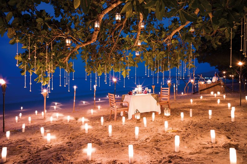 Romantisches Stranddinner Ein romantisches Abendessen am Strand bei Nacht, umgeben von zahlreichen Kerzen und unter einem Baum, von dem hängende Lichter dekoriert sind.
