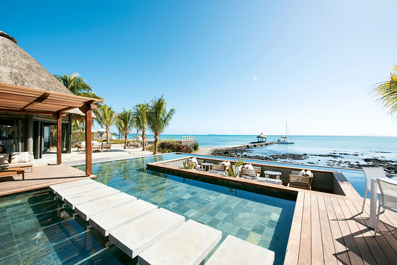 Außenareal Luxuriöses Resort mit Infinity-Pool und Holzdeck, vorne Trittsteine im Wasser und ein Strohdachhaus links, inklusive Strandblick mit Palmen und einem Segelboot am Horizont unter blauem Himmel.