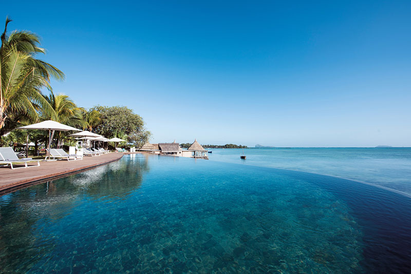 Infinitypool Ein luxuriöses Infinity-Pool am Strand mit klarem blauem Wasser, umgeben von Palmen und Sonnenliegen. Im Hintergrund sind traditionelle Strohdachbauten und das ruhige Meer unter einem strahlend blauen Himmel zu sehen.