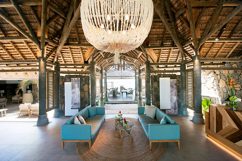 Lobby Luxuriöse Lounge mit rustikalem Charme und offener Architektur, ausgestattet mit einer großen Hängeleuchte aus Perlenschnüren, eleganten blauen Sofas und einem Naturholztisch auf einer runden Matte. Der Raum ist von Steinpfeilern umgeben und zeigt eine Decke aus traditionellem strohartigem Material, was eine einladende, tropische Atmosphäre schafft.