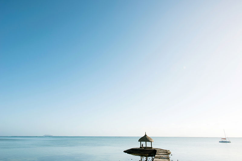 Meerblick Ein friedvolles maritimes Panorama mit einem Pavillon, der auf einem kleinen Vorsprung nahe der Wasserlinie steht, während im Hintergrund eine ruhige See und ein schmales Boot zu sehen sind. Der klare Himmel und das ruhige Meer erzeugen ein Gefühl von Weite und Stille.