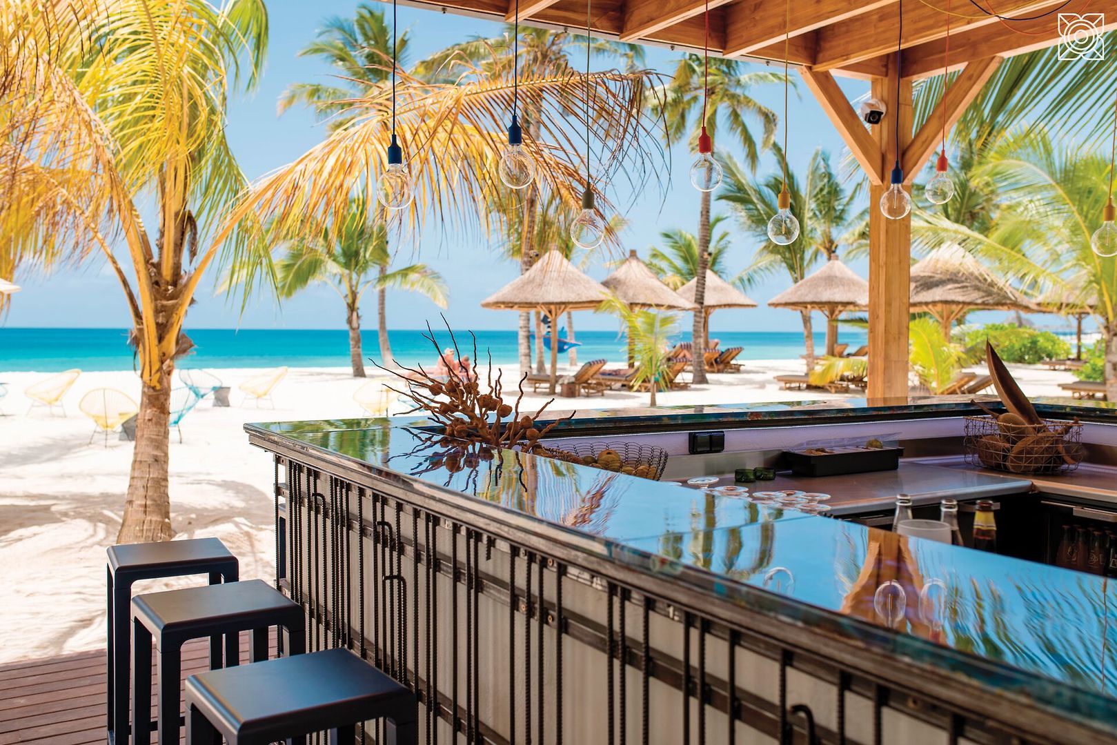 Bahari Bar Blick von einer Strandbar mit glänzender Theke und Barhockern auf einen tropischen weißen Sandstrand mit Palmen und Strohschirmen unter einem klaren blauen Himmel.
