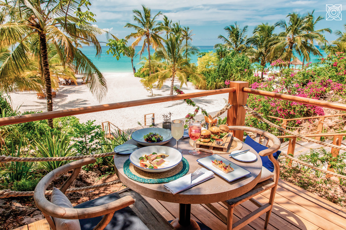 Köstliche Speisen mit Meerblick Blick auf einen gedeckten Tisch mit frischen Mahlzeiten auf einer Terrasse mit Blick auf einen tropischen Strand mit klarem blauen Wasser und Palmen.