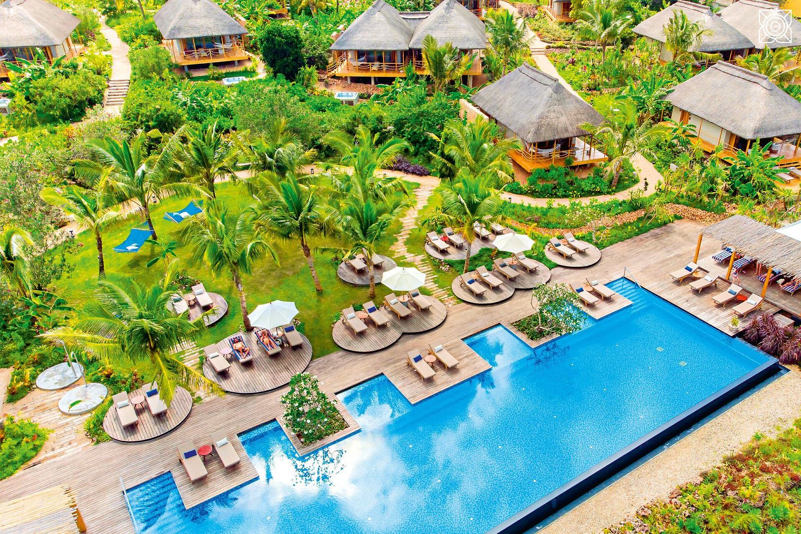Infinitypool Ein luxuriöses Ferienresort mit eingebetteten Bungalows mit Reetdach umgeben von üppigem Grün. Im Vordergrund ein großer Swimmingpool flankiert von Holzliegen und weißen Sonnenschirmen.