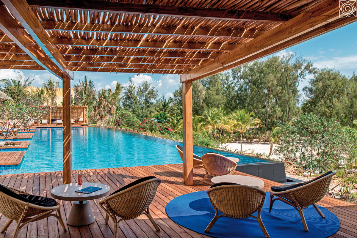 Maisha Resto & Bar Ein luxuriöses Außenterrassen-Setting mit einer Holzpergola und darunter modernen Rattanmöbeln, welche einen entspannenden Blick auf einen langgezogenen blauen Infinity-Pool bieten, der in eine üppige, tropische Landschaft mit hohen Palmen und dichtem Grün übergeht. Ein idyllisches Urlaubsszenario mit klarem Himmel im Hintergrund.