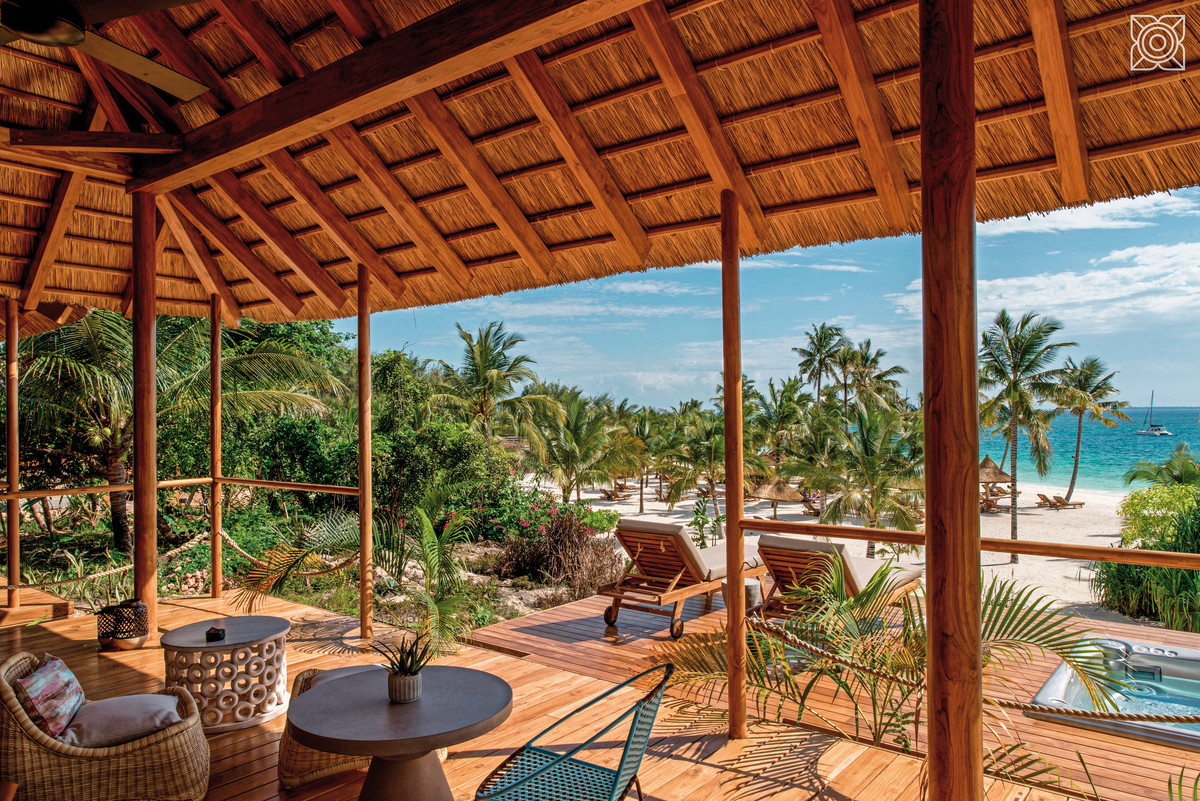 Terrasse der Ocean Front Suite Blick von einer Holzterrasse mit strohgedecktem Dach und entspannenden Sitzmöbeln auf einen tropischen Strand mit Palmen und klarem Himmel. Im Hintergrund ist das ruhige Meer mit einem segelnden Boot zu sehen.