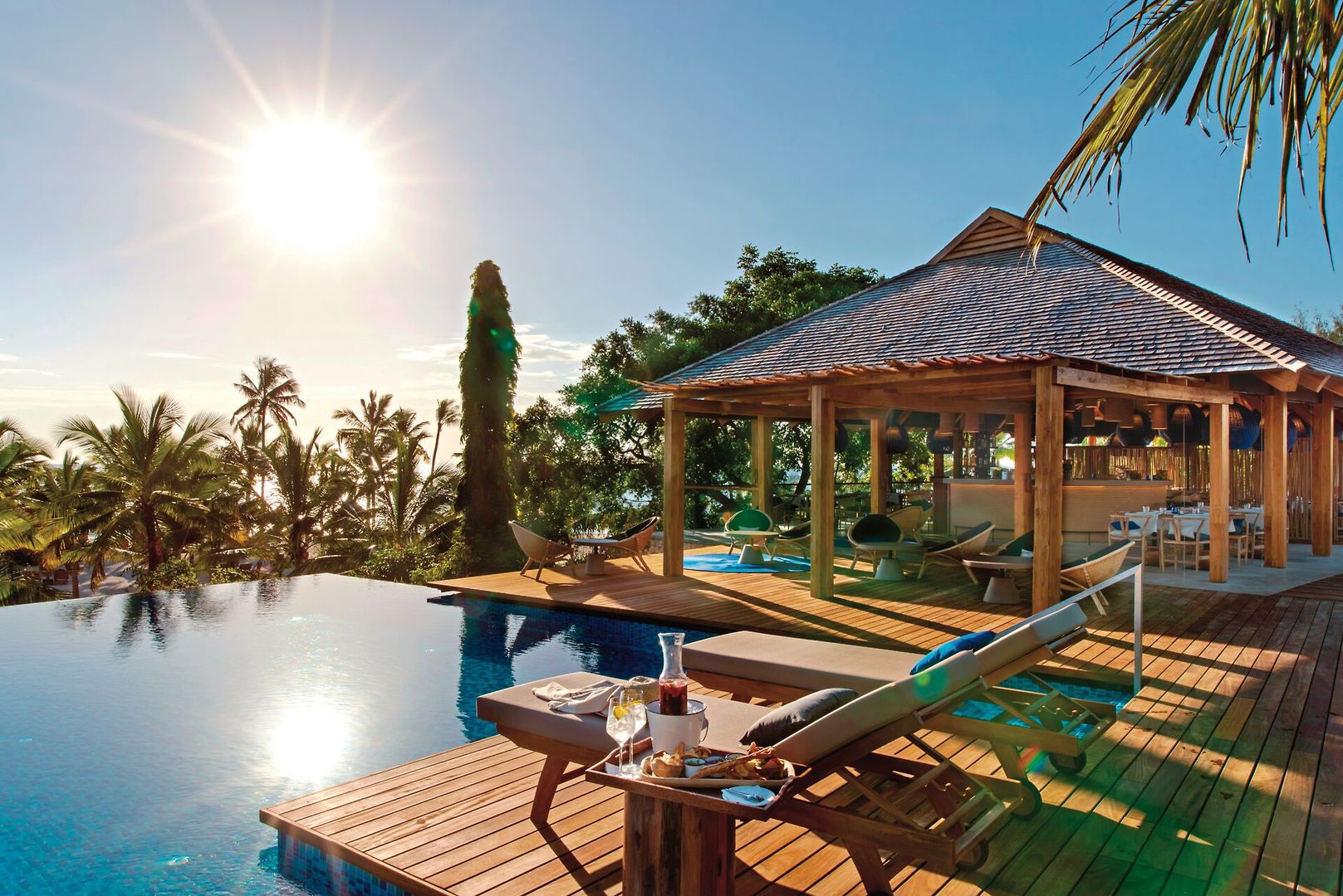 Swimmingpool Ein luxuriöses Urlaubsresort mit Infinity-Pool, Holzliegestühlen und einem gedeckten Tisch mit einem Blick auf tropische Bäume unter einem klaren blauen Himmel mit strahlender Sonne. In der Nähe des Pools befindet sich ein offenes Pavillon mit Sitzbereichen.