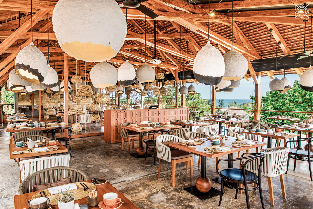 Upendo Restaurant Das Bild zeigt den Innenbereich eines stilvollen Restaurants mit einer rustikalen Holzdecke und offenem Raumkonzept. Über den sorgfältig gedeckten Tischen hängen große, kreative Leuchten in Tropfenform, die aus natürlichen Materialien wie Bambus und Rattan zu bestehen scheinen. Durch die große, offene Fensterfront im Hintergrund ist ein Blick auf das Meer möglich, was auf eine Küstenlage hindeutet. Der Raum strahlt eine warme und einladende Atmosphäre aus.