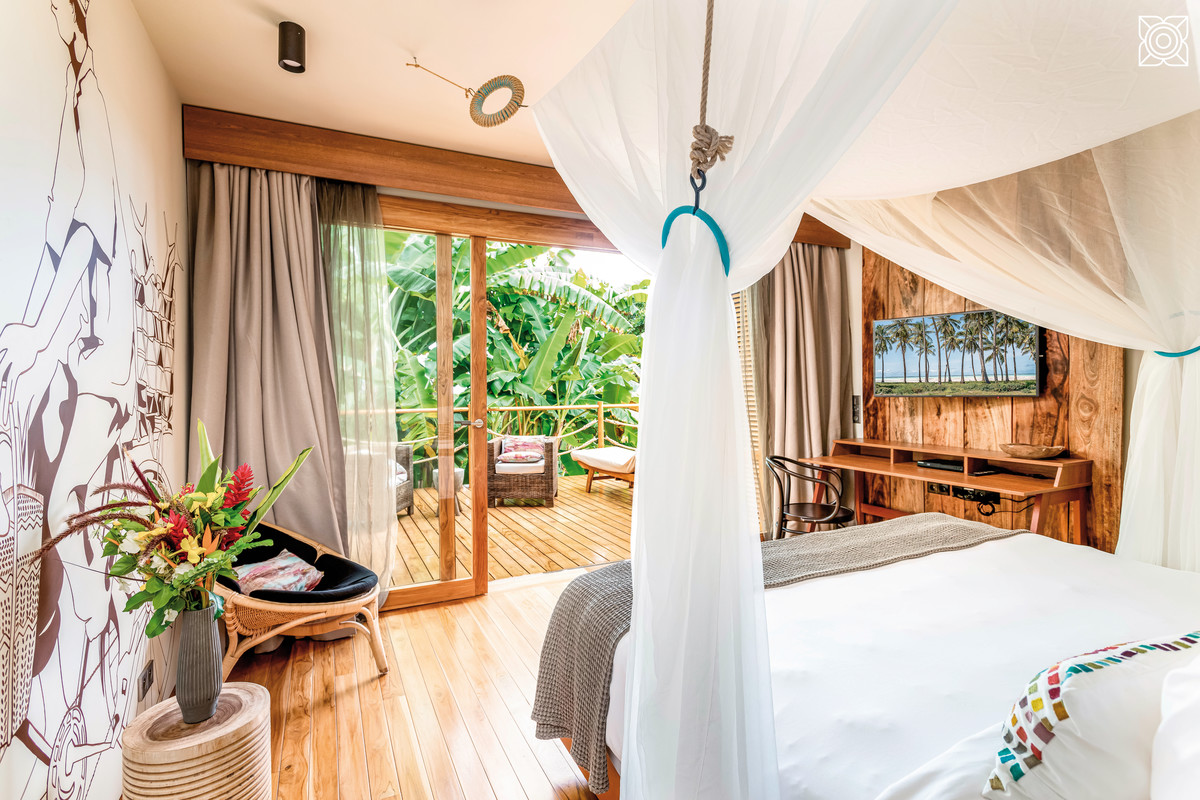 Wohnbeispiel Ein gemütliches und stilvolles Schlafzimmer im tropischen Ambiente mit großem, weißem Himmelbett und Moskitonetz. Die Schiebetür öffnet sich zu einer Holzterrasse mit Blick auf üppige Vegetation. Die Wände sind mit einem großen Wandbild verziert, und die Einrichtung umfasst eine Schreibkonsole mit Stuhl und eine dekorative Vase mit frischen Blumen.