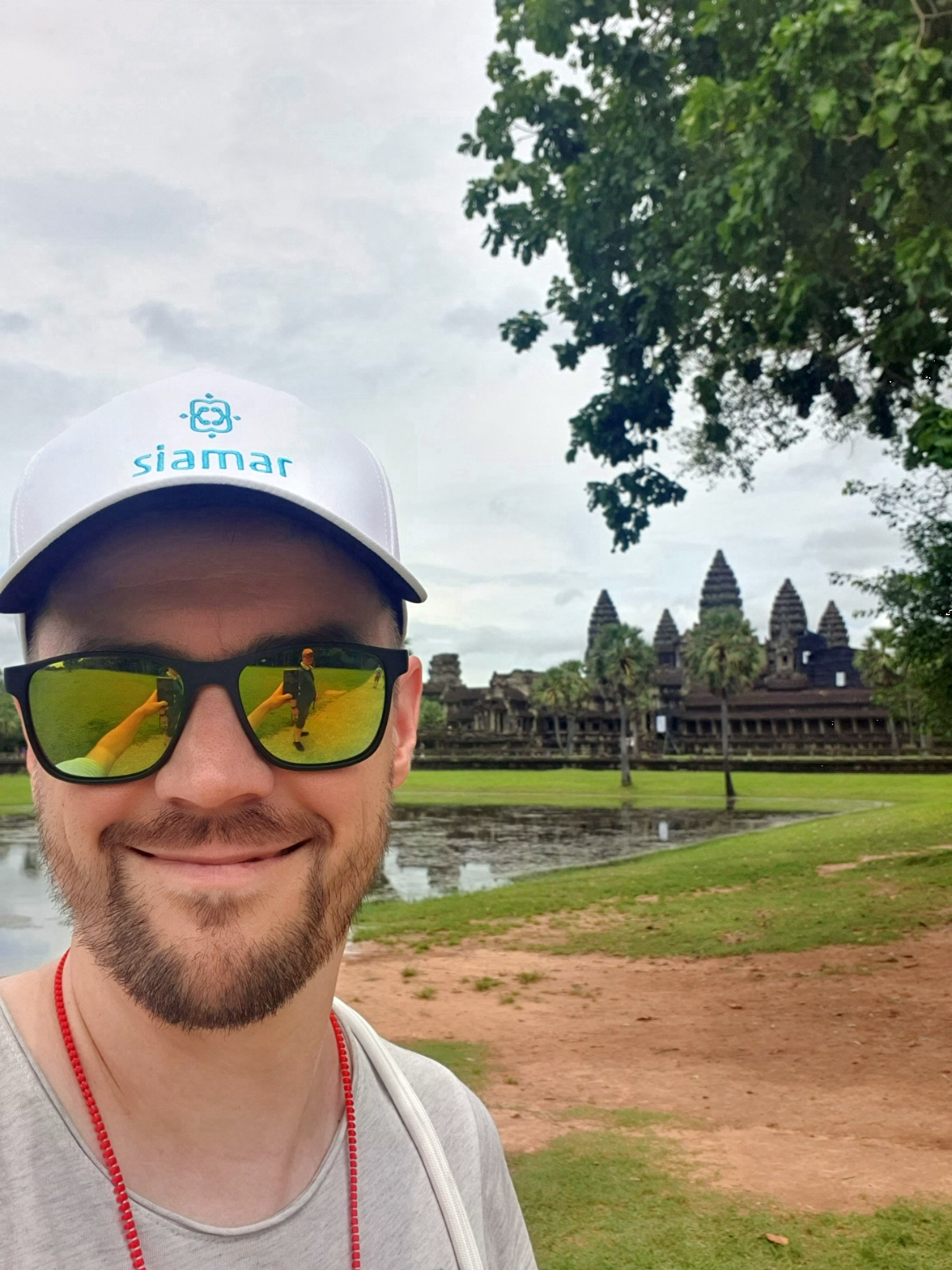 Christoph Busch in Kambodscha Lächelnder Mann mit Sonnenbrille und weißer Schirmmütze macht ein Selfie mit der Tempelanlage Angkor Wat im Hintergrund. Es ist leicht bewölkt und die Umgebung ist grün mit sichtbaren Wasserflächen.