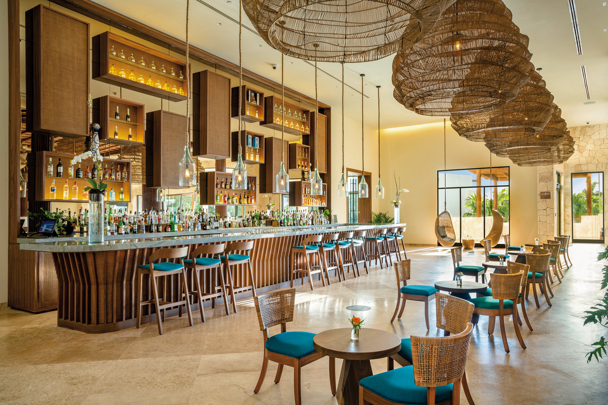 Bar Ein stilvoll eingerichteter Hotelbar-Bereich mit einer Bartheke, an der blaue Barhocker stehen. Über der Theke schweben große, korbartige Hängeleuchten. Flaschen und Gläser sind in Regalen an der Wand ausgestellt. Vor der Theke laden kleine Tische mit gewebten Stühlen zum Verweilen ein, während Tageslicht den Raum durch große Fenster auf der rechten Seite erhellt.