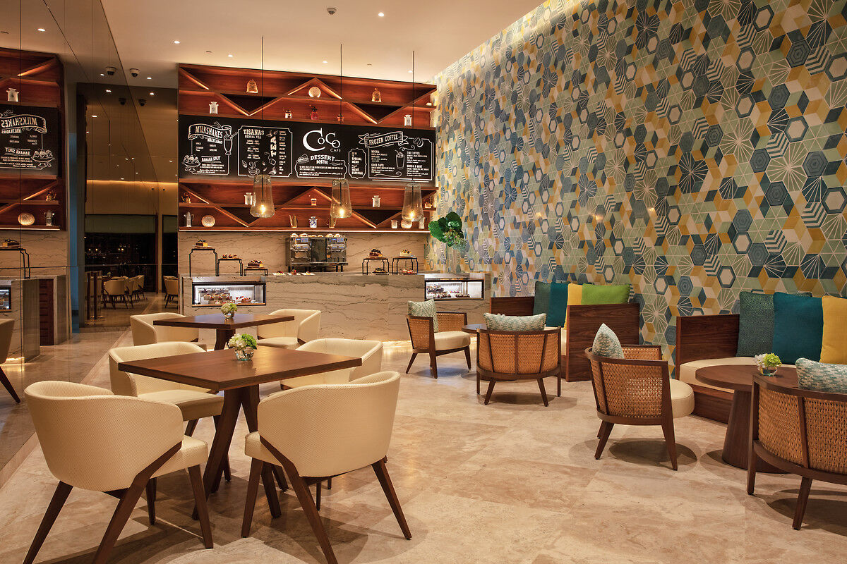 Coco Café Ein modernes Café mit stilvoller Inneneinrichtung. Im Hintergrund sind eine beleuchtete Menütafel und eine Kaffeemaschine auf einem Marmortheke zu sehen. Im Vordergrund stehen elegante Tische und Stühle mit bequemen Polstern. An der rechten Wand fällt ein Muster aus geometrischen Formen in verschiedenen Blautönen auf. Die Atmosphäre wirkt einladend und zeitgemäß.