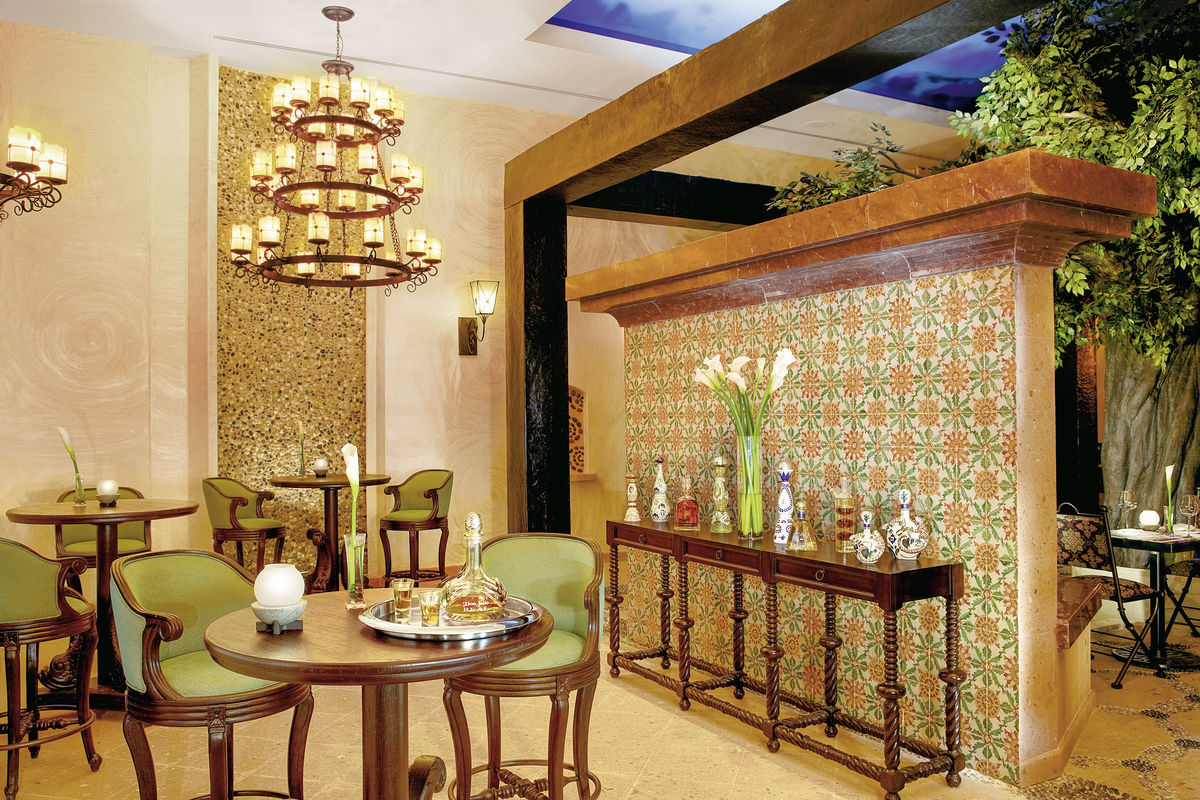 El Patio Restaurant Ein luxuriös eingerichtetes Zimmer im marokkanischen Stil mit gemusterten Fliesen an der Wand und einer stilvollen, dunkelholzigen Bartheke. Zwei Kronleuchter erhellen das Zimmer, das zusätzlich mit grünen Polsterstühlen und runden Holztischen ausgestattet ist. Dekorative Gegenstände wie Vasen und Flaschen schmücken die Thekenoberfläche, während Pflanzen im Hintergrund das Ambiente ergänzen.
