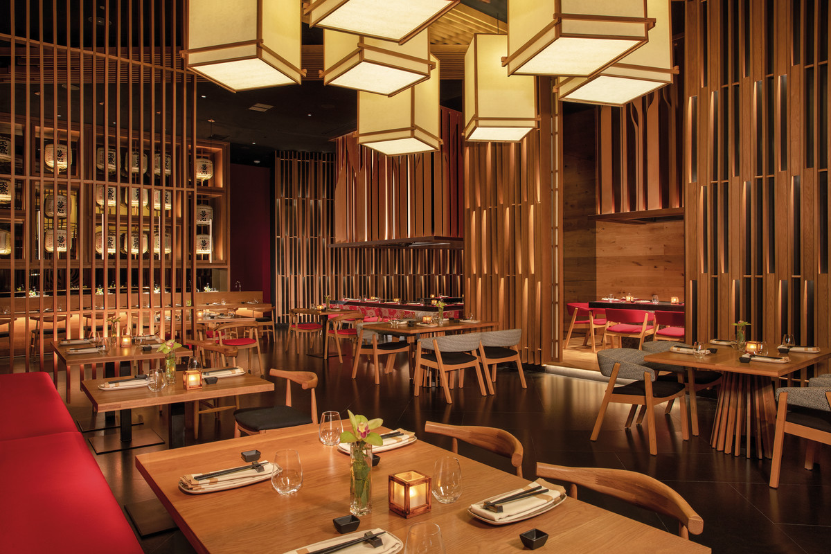 Himitsu Restaurant Innenansicht eines stilvoll eingerichteten Restaurants mit asiatischer Ästhetik. Holzvertäfelungen und -säulen strukturieren den Raum, während quadratische Hängelampen warmes Licht spenden. Mehrere elegant gedeckte Tische mit hölzernen Stühlen und roten Akzenten laden zum Verweilen ein. Ein Teil der Wand ist durch ein großes Regal mit asiatischen Dekorationsgegenständen ausgefüllt.