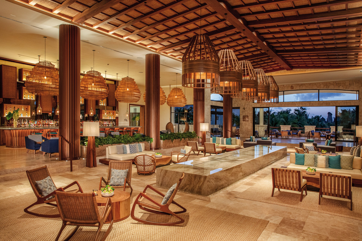 Lobby Ein luxuriöser Hotel-Lobbybereich mit hohen Decken und Holzbalken. Mehrere Hängeleuchten in Korbgeflechtoptik beleuchten den Raum. Eine Bar befindet sich im Hintergrund, umgeben von blauen Barhockern, während im Vordergrund elegante Lounge-Möblierung mit Tischen und bequemen Stühlen angeordnet ist. Der Boden ist mit großflächigen Natursteinfliesen ausgelegt, ergänzt durch dekorative Teppiche. Durch die großen Fensterfronten ist ein Blick auf Palmen und eine Dämmerungsszenerie möglich.
