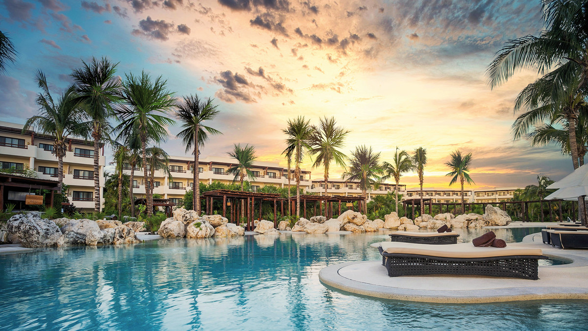 Pool Ein luxuriöser Resort-Poolbereich bei Sonnenuntergang mit Liegestühlen auf einer kleinen Insel im Wasser, umgeben von Palmen und einem ansprechenden Hotel im Hintergrund.