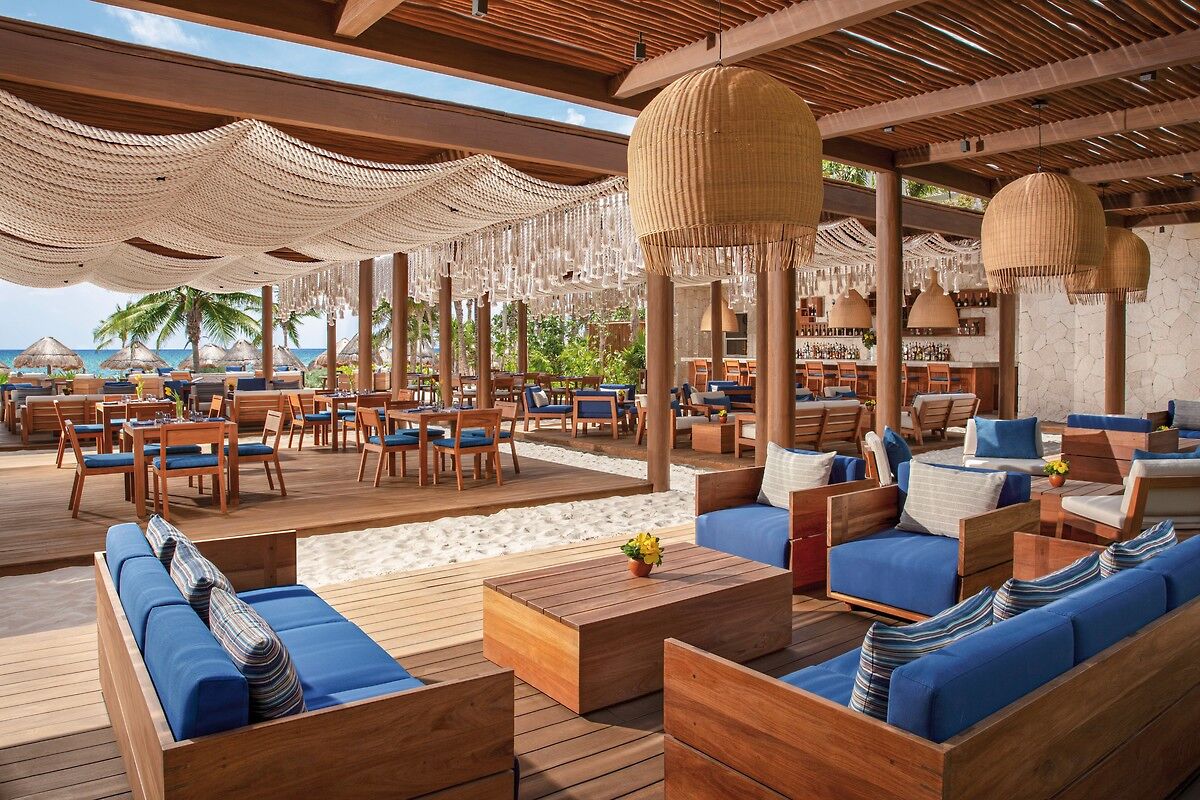 Preferred Beach Club Ein stilvoller, überdachter Strandbereich mit eleganter Inneneinrichtung: hängende Kugelleuchten aus Rattan, gewebte Decken, Holzmöbel mit blauen Polstern und Tischen. Im Hintergrund sind Palmen und ein sonniger Himmel zu erkennen.