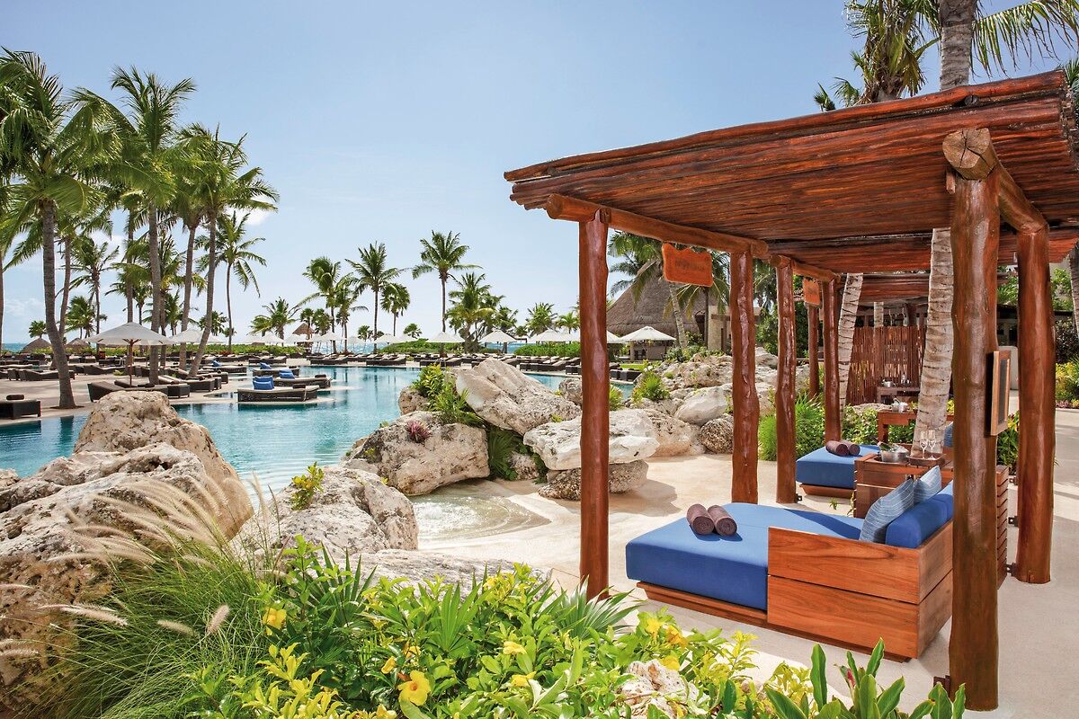 Sonnenliegen am Pool Ein luxuriöser Außenbereich eines Resorts mit einem ruhigen, von Palmen gesäumten Pool und eleganten Liegestühlen. Vor im Bild befindet sich ein Holzpavillon mit bequemen Liegen und einer Aussicht auf den Pool- und Gartenbereich.