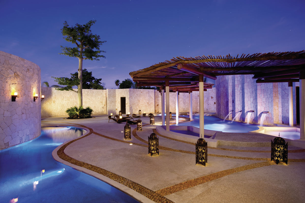 Spa Ein luxuriöses Spa mit beleuchtetem Pool bei Dämmerung, umgeben von hohen Mauern und einer Pergola, unter einem sternenklaren Himmel. Dekorative Laternen verteilen sich auf dem eleganten Bodenbelag und sorgen für ein warmes, einladendes Ambiente.