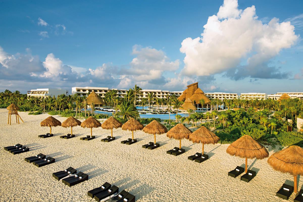 Strand Bild eines luxuriösen Strandresorts mit Reetdachhütten, Sonnenliegen, üppiger Vegetation und modernen Gebäuden im Hintergrund unter einem blauen Himmel mit flauschigen Wolken.