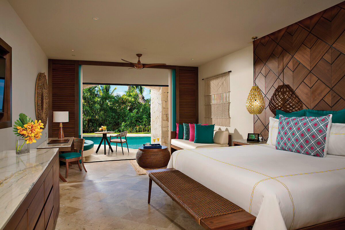 Wohnbeispiel Ein luxuriös eingerichtetes Hotelzimmer mit großen Fenstern, durch die man auf eine Terrasse mit Blick auf einen privaten Pool und tropische Vegetation sieht. Das Zimmer ist mit einem großen Bett mit bunten Kissen, einer Wand mit Holzverkleidung, einer Decke mit Ventilator und stilvollen Dekorationselementen wie einem Flechtbank und Hängeleuchten ausgestattet. Im Vordergrund ist eine elegante Marmoroberfläche mit frischen Blumen zu sehen.