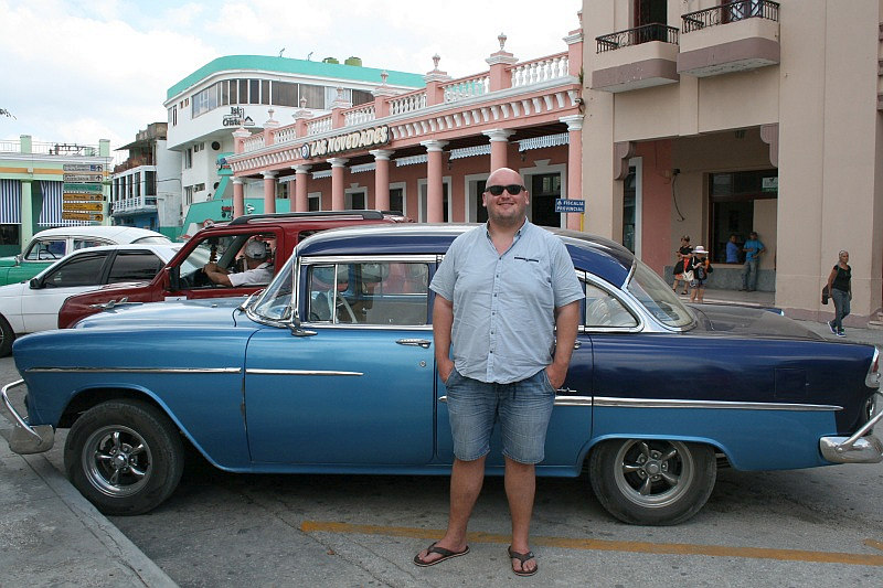 Oldtimer allerorten – auch in Holguin Ein Mann steht lächelnd vor einem blau-weißen klassischen Auto auf einer Straße mit pastellfarbenen Gebäuden im Hintergrund.