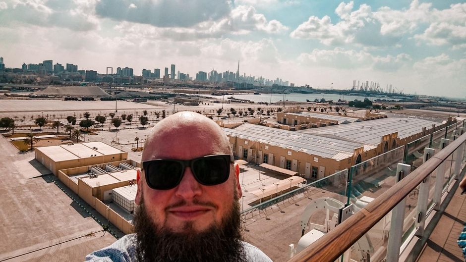 Markus Patho in Dubai Ein Mann mit Bart und Sonnenbrille macht ein Selfie auf einer Terrasse mit Blick auf eine urbane Skyline im Hintergrund. Niedrige Gebäude befinden sich im Vordergrund, während im Hintergrund eine moderne Stadt mit Wolkenkratzern unter einem blauen, leicht bewölkten Himmel zu sehen ist.