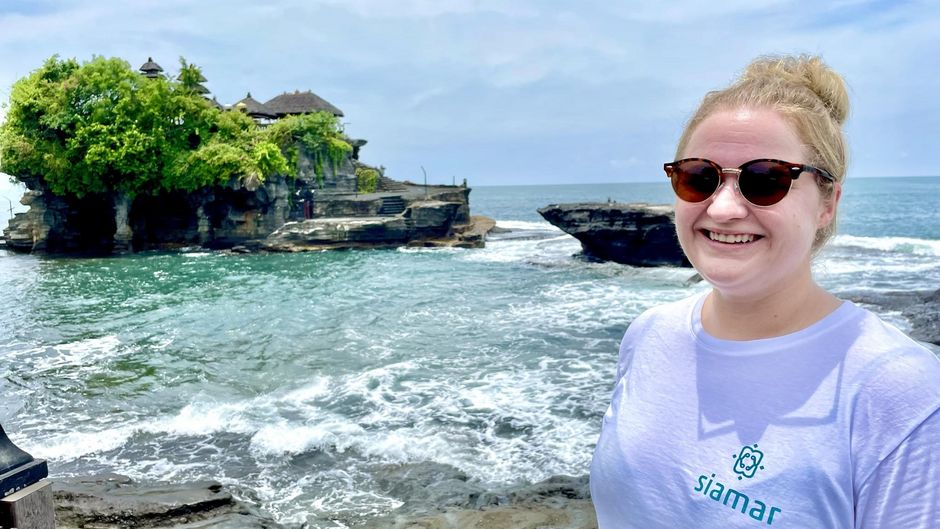 Franziska Martin vor Tanah Lot Eine fröhliche Person mit Sonnenbrille lächelt in die Kamera vor dem Hintergrund eines beeindruckenden Tempels auf einer kleinen Felsinsel, umgeben vom rauschenden Meer.