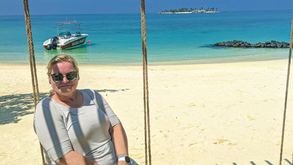 Meike Schulze schaukelt Eine Person mit Sonnenbrille sitzt entspannt auf einer Schaukel am Strand, im Hintergrund sind klares türkisfarbenes Wasser, ein Motorboot und eine kleine Insel mit Palmen zu sehen.