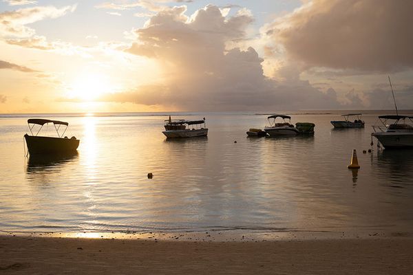 Boote im Sonnenuntergang Alternative Text: Mehrere Boote liegen bei Sonnenuntergang in ruhigem Wasser nahe einem Strand mit sanften Reflexionen der abnehmenden Sonne im Wasser.