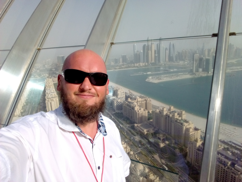 Markus Patho auf dem Aussichtspunkt The View Ein Mann mit Glatze und Sonnenbrille macht ein Selfie vor dem Panoramablick auf eine Küstenstadt mit Hochhäusern, vermutlich von einem hohen Aussichtspunkt aus gesehen. Er trägt ein weißes Hemd mit aufgekrempelten Ärmeln und freut sich offensichtlich über den Ausblick. Die Sonne scheint, und die Sicht ist leicht dunstig.