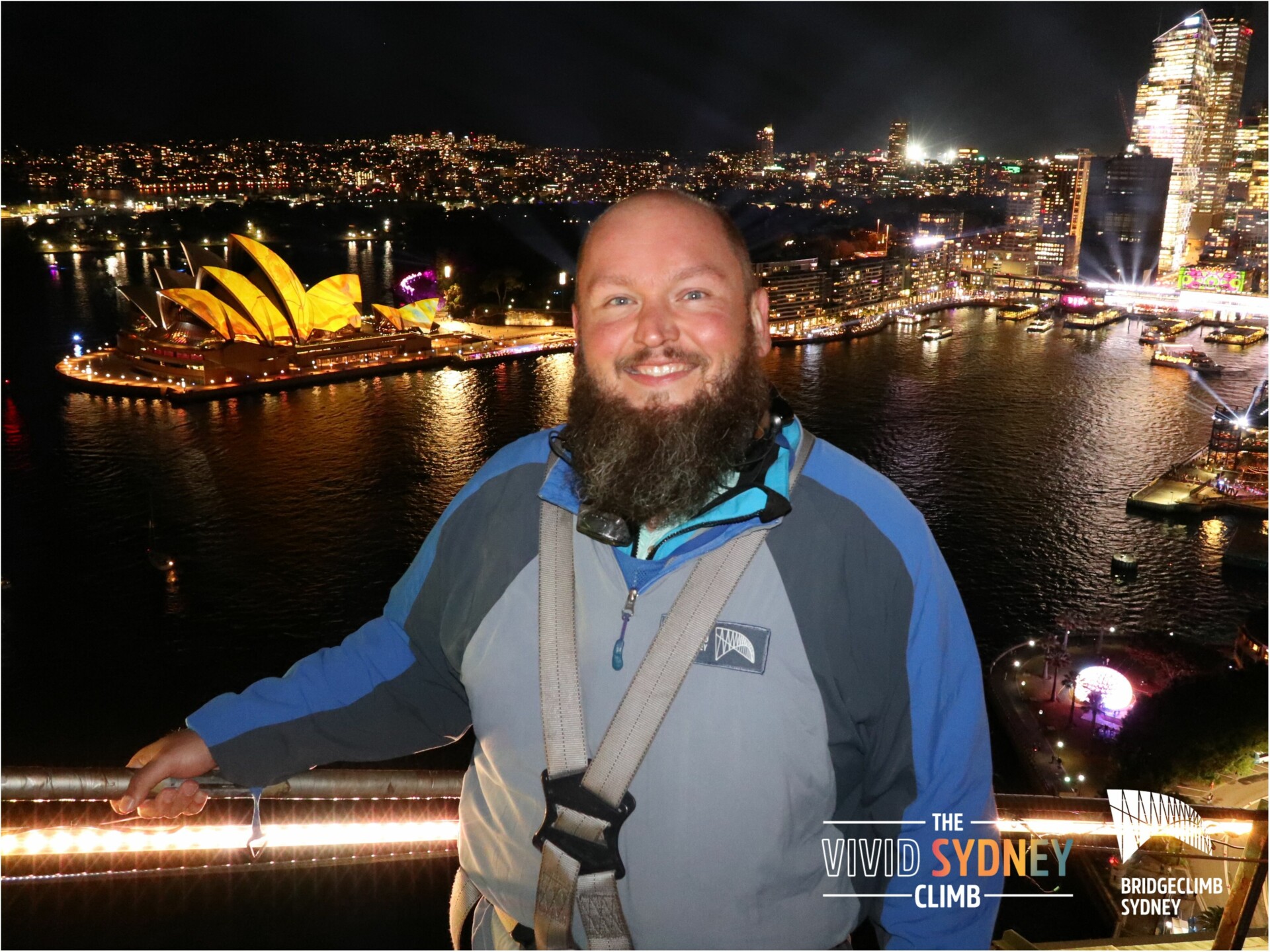 Markus Patho vor der Oper in Sydney Ein lächelnder Mann mit Bart in Sicherheitsausrüstung steht auf einer Aussichtsplattform bei Nacht. Im Hintergrund ist die beleuchtete Skyline von Sydney mit dem markanten Sydney Opera House und der Hafenbrücke zu sehen. Lichter reflektieren im Wasser des Hafens, und die Stadt wirkt lebhaft beleuchtet. Textelemente im Bild weisen auf "The Vivid Sydney Climb" und "Bridgeclimb Sydney" hin.